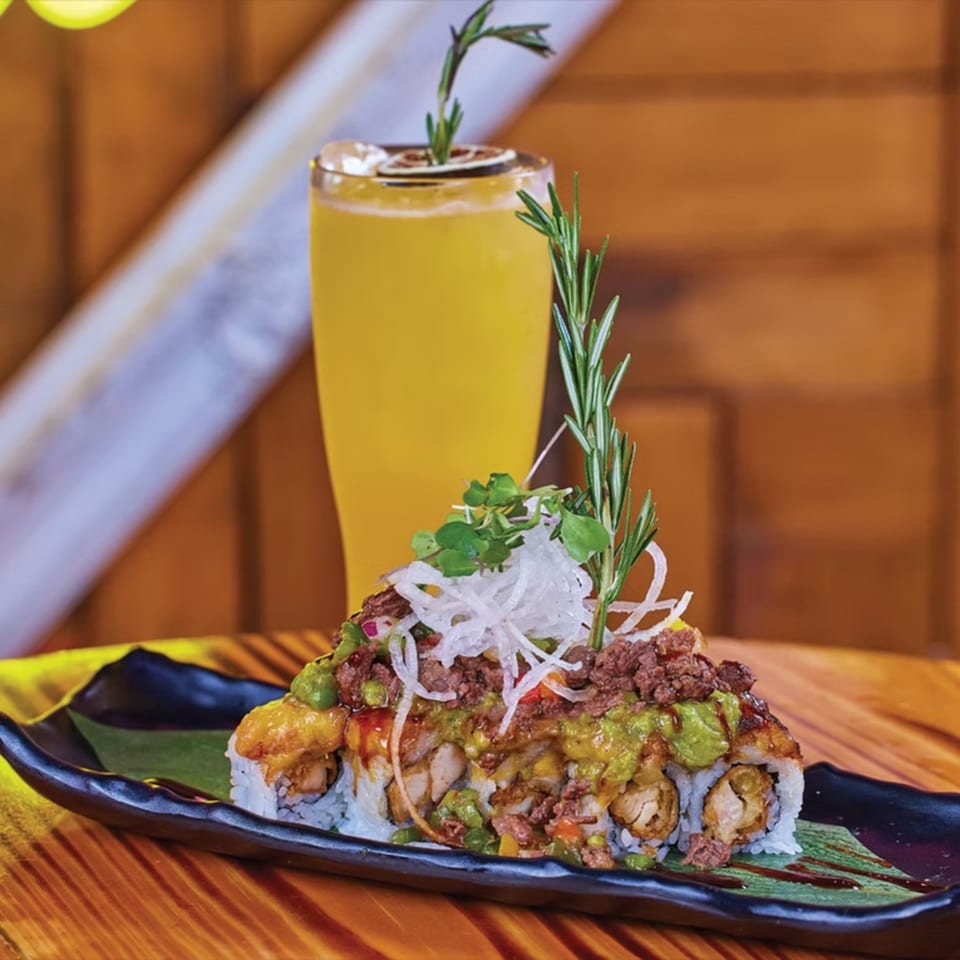 El Tiesto Cafe's sushi roll topped with meat, avocado, and onions, with a drink in the background.