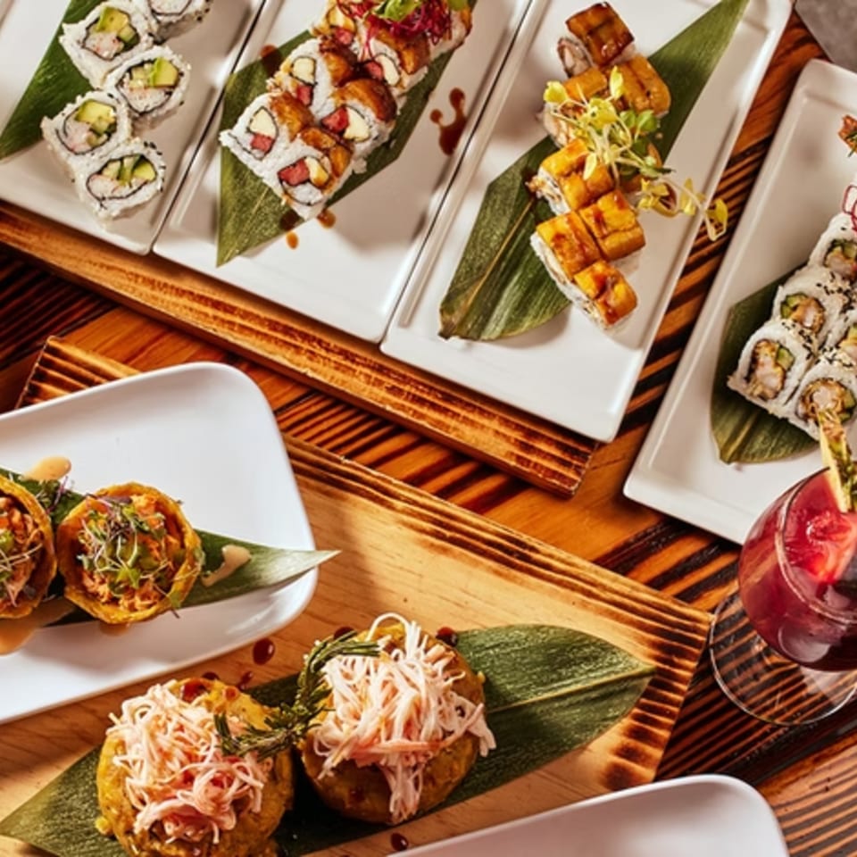Assortment of sushi rolls, plantain cups, and a red cocktail on a wooden table.