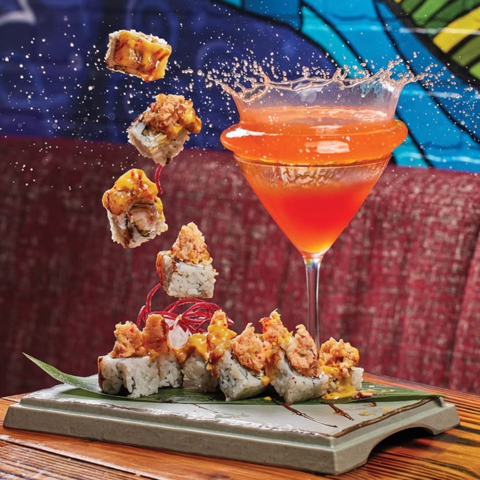 Sushi rolls with toppings and a splashing orange cocktail against a colorful background.