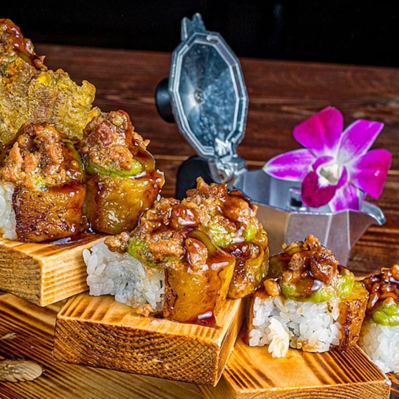 Sushi rolls with green filling and brown topping, served on wooden blocks with a purple orchid.