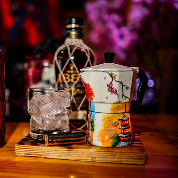 A decorative moka pot, a glass of ice, and a bottle of rum on a wooden surface.