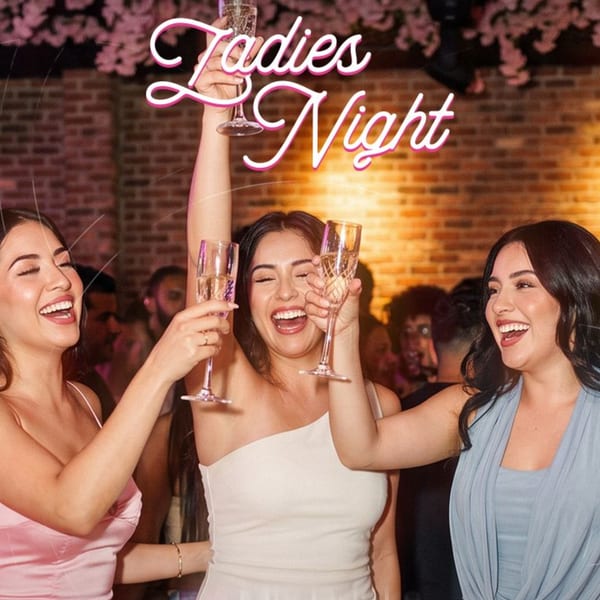 Three women laugh and raise champagne glasses during a lively restaurant gathering beneath a “Ladies Night” sign.