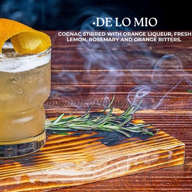 De Lo Mio cocktail with orange peel, rosemary, and smoke on a wooden board.