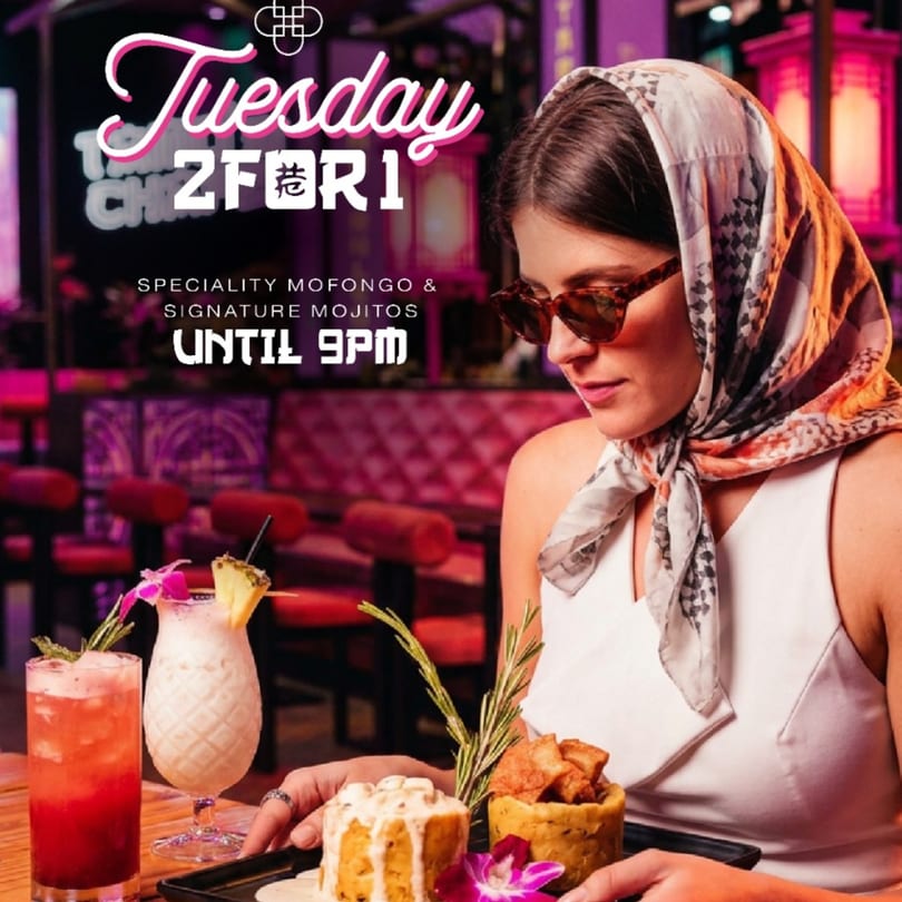 Woman in sunglasses and headscarf holding a tray with mofongo dishes and cocktails in a vibrant restaurant.