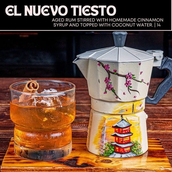 A cocktail with ice and a cinnamon stick next to a decorative coffee pot on a wooden surface.