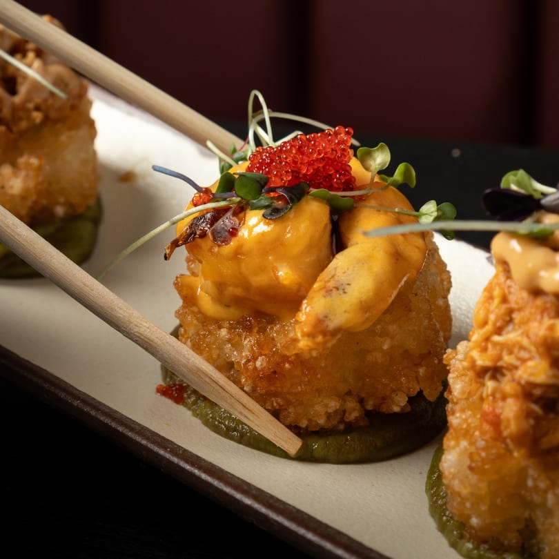 Crispy fried sushi rolls topped with orange sauce, red caviar, and microgreens, with chopsticks.