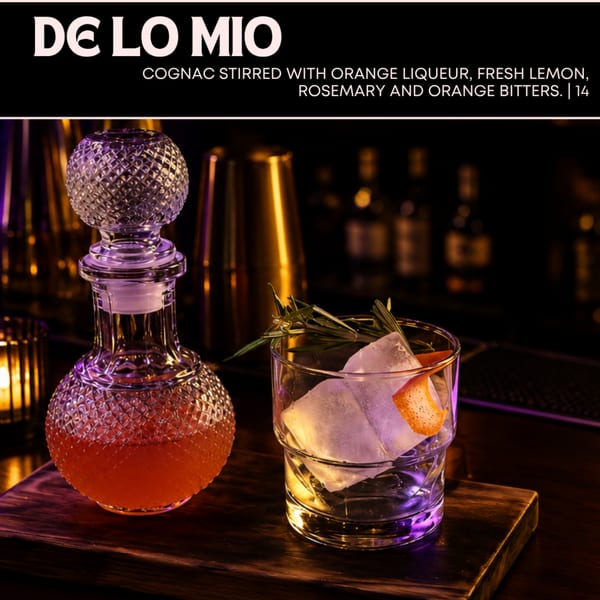A De Lo Mio cocktail with ice, rosemary, and orange peel, next to a crystal decanter.
