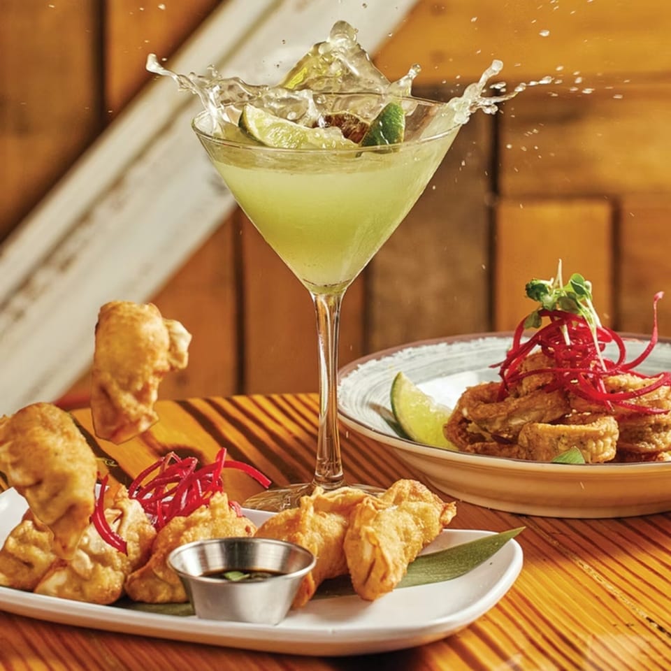 A lime green cocktail splashes in a martini glass next to fried dumplings and calamari.