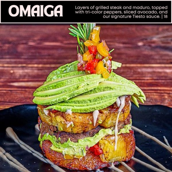 A tall OMAIGA steak and maduro dish, topped with peppers, avocado, and Tiesto sauce.