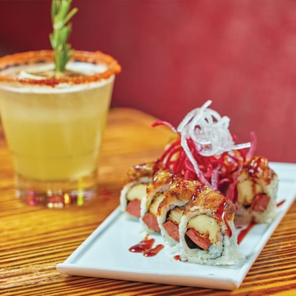 Sushi rolls with hot dog and cheese, topped with sauce and shredded vegetables, next to a cocktail.
