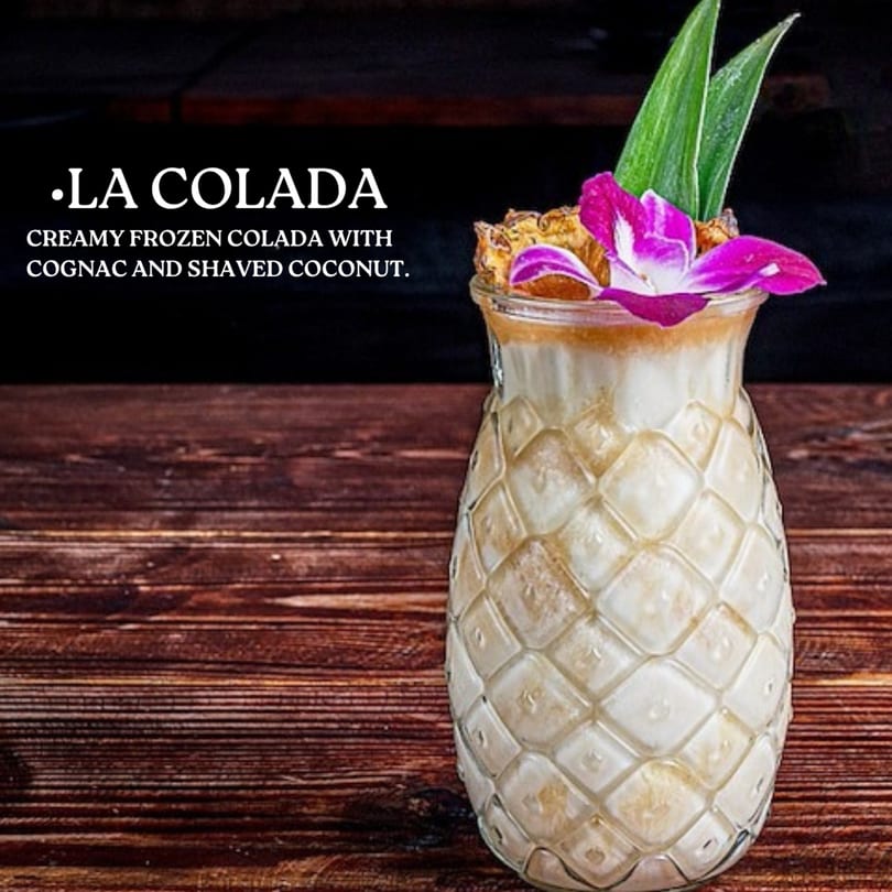 A La Colada cocktail in a pineapple-shaped glass, garnished with a purple orchid and green leaves.