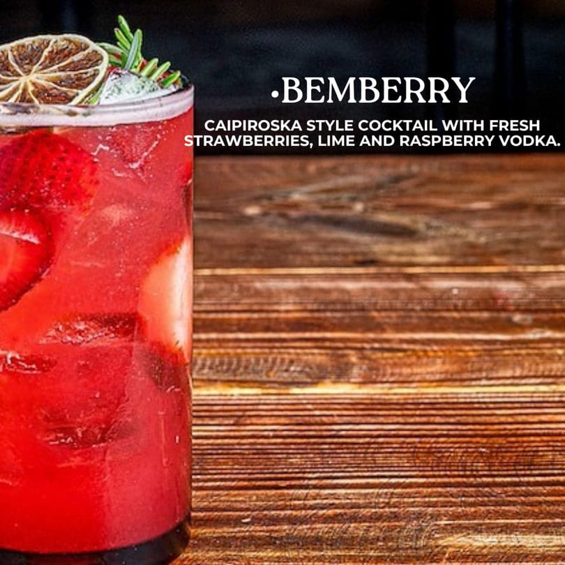 A vibrant red cocktail with strawberries, garnished with a dried lime slice and rosemary sprigs.