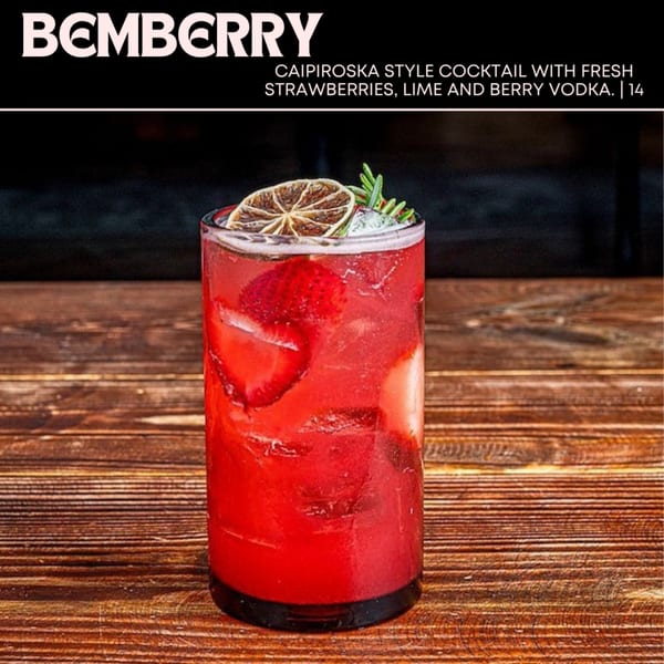 A vibrant red Bemberry cocktail with strawberries, lime, and rosemary on a wooden table.